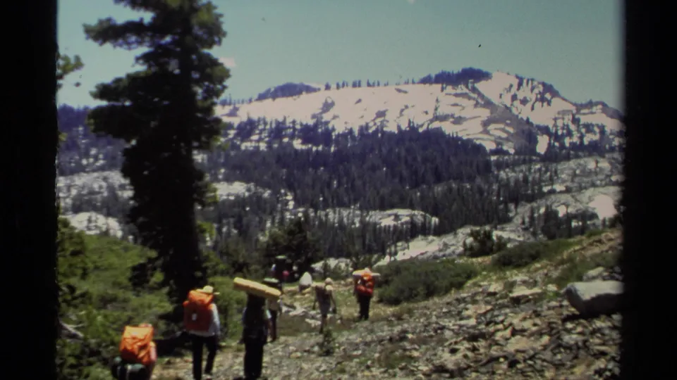 1978 Lake Tahoe 8mm frame showing backpackers walking across a rocky hillside with pine trees and snowy alpine ridges behind them.