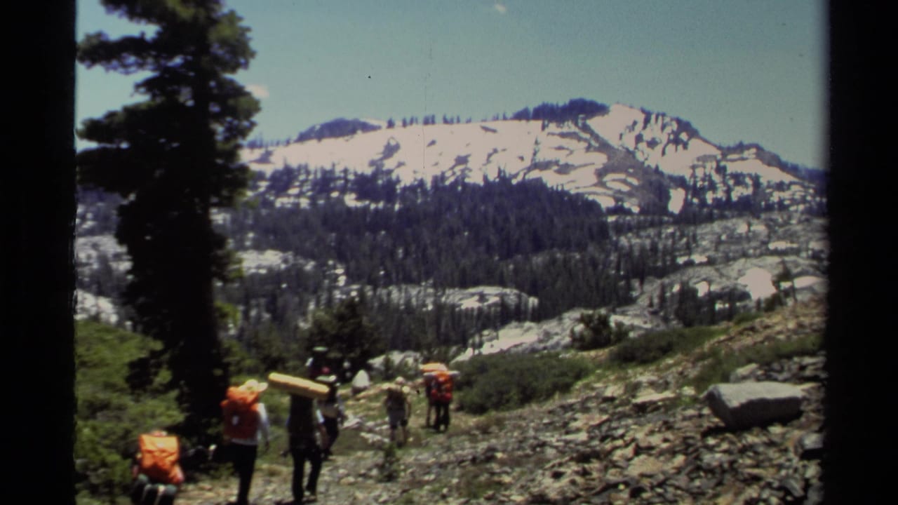 Hikers with backpacks descend a rocky mountain trail below snowy peaks and pine trees.