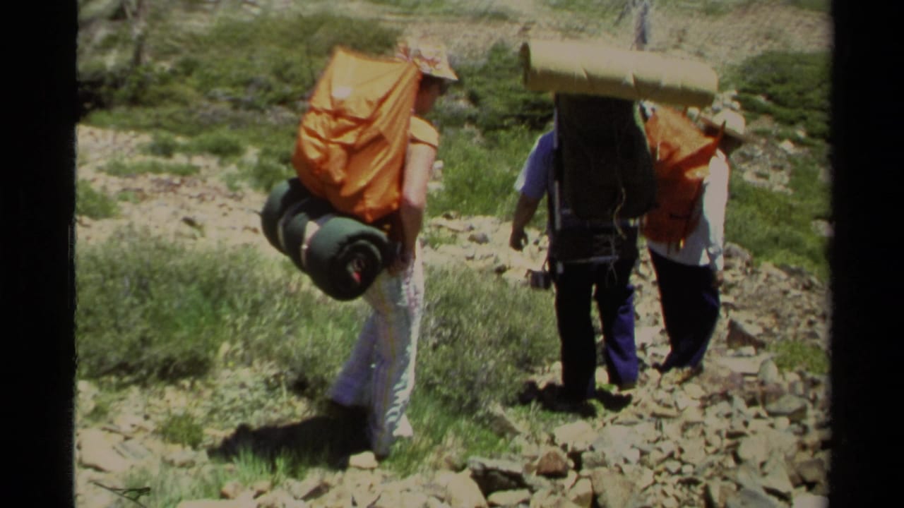 Three backpackers walk away on a rocky trail through green mountain terrain.
