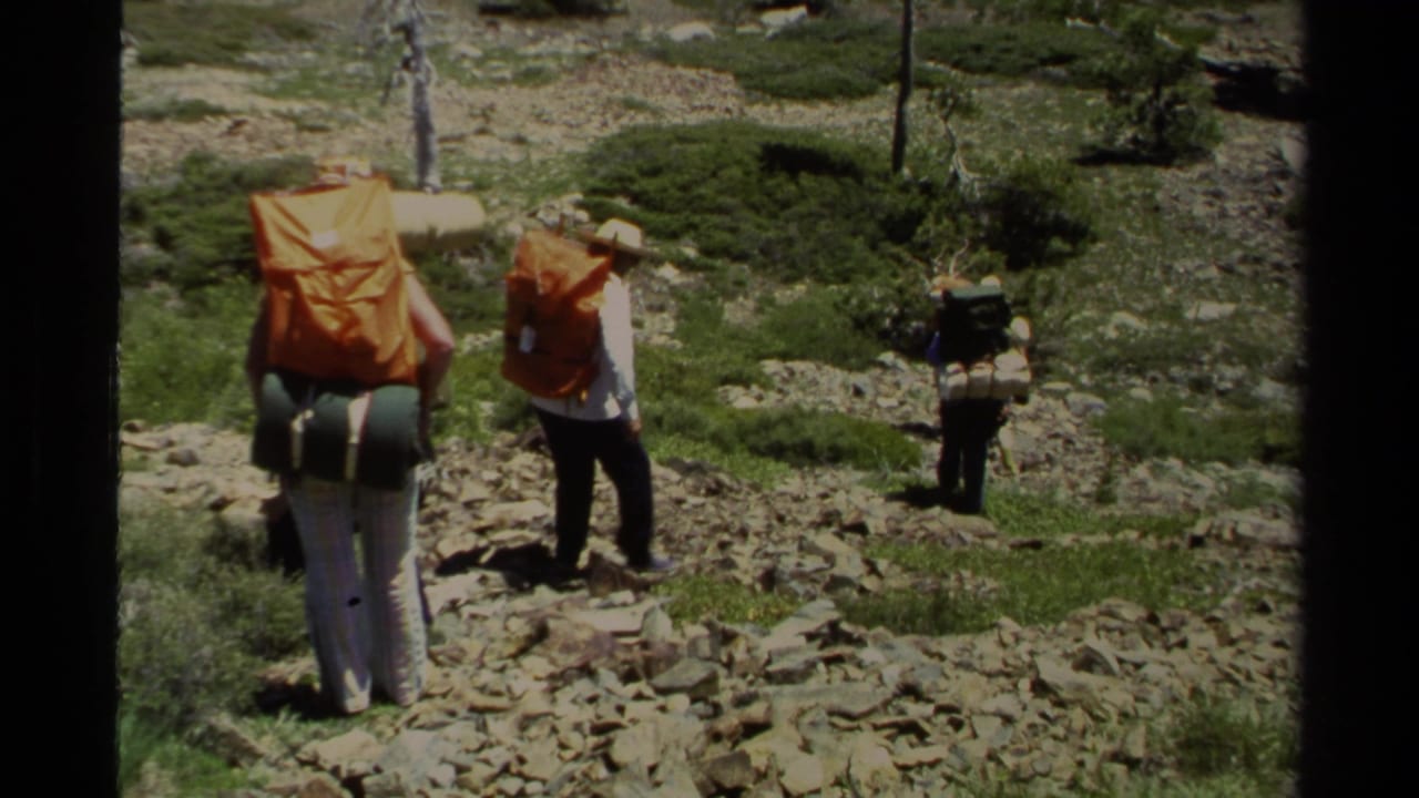 1978 Lake Tahoe home-movie frame of three hikers carrying large orange and green packs across a rocky mountain trail.