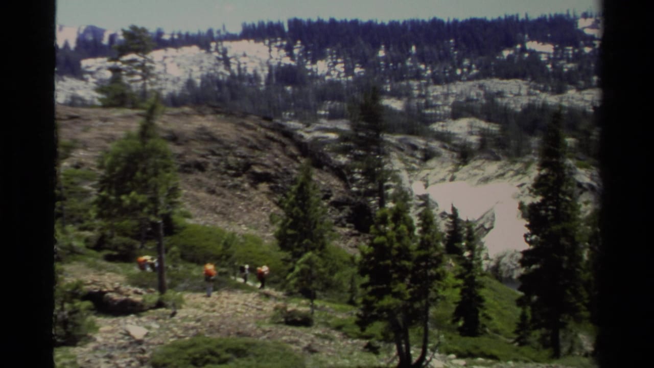Wide 1978 Lake Tahoe view with hikers crossing a rocky slope, evergreen trees, and snow patches in the distance.