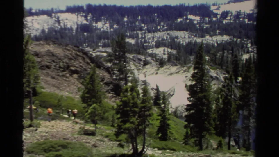 1978 Lake Tahoe footage frame showing backpackers on a rocky incline beneath evergreen trees and distant snow.