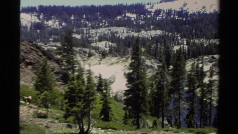 Wide Lake Tahoe mountain view from 1978 with evergreen trees, distant backpackers, rocky slopes, and lingering snow.