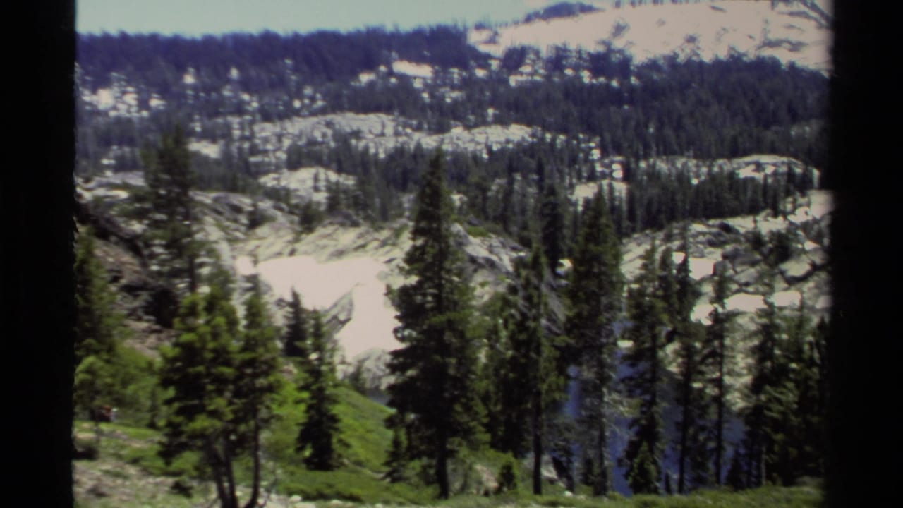 1978 Lake Tahoe home-movie frame showing snow patches, rocky terrain, dense evergreens, and a green foreground slope.