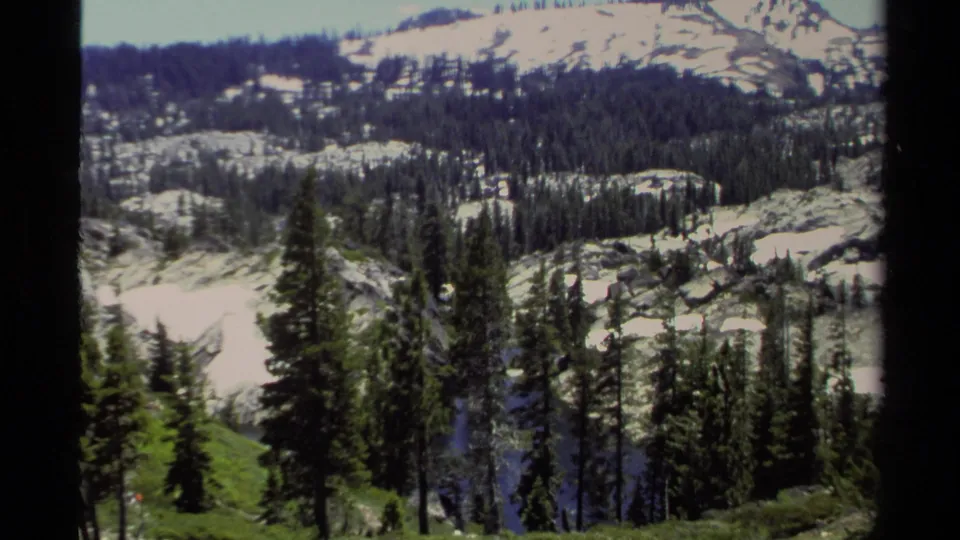 1978 8mm view of Lake Tahoe mountain terrain with evergreen forest, lingering snow, and a green slope in the foreground.