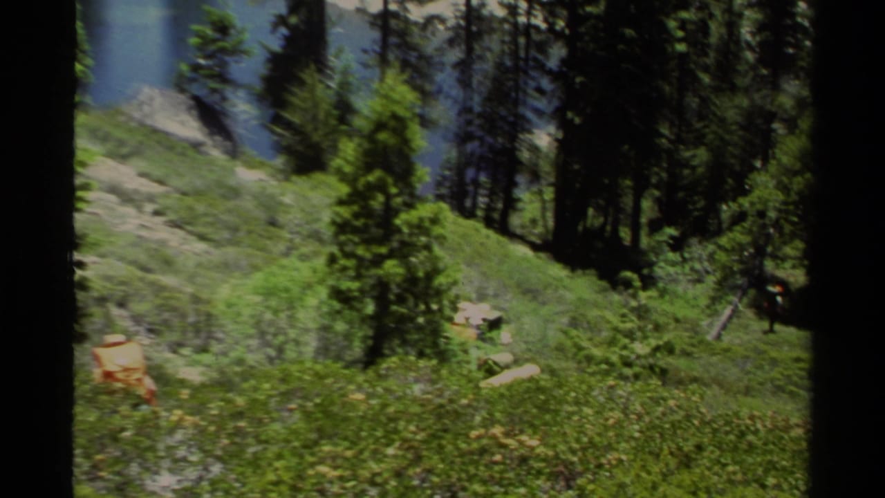 1978 Lake Tahoe home-movie frame showing hikers with packs on a green hillside trail among pines, with lake water partly visible.