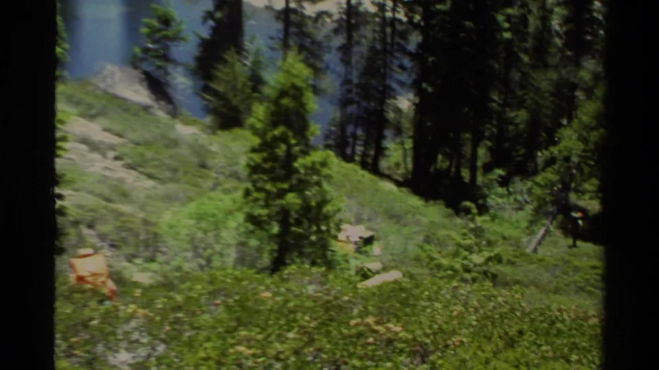 1978 Lake Tahoe home-movie frame showing hikers with packs on a green hillside trail among pines, with lake water partly visible.