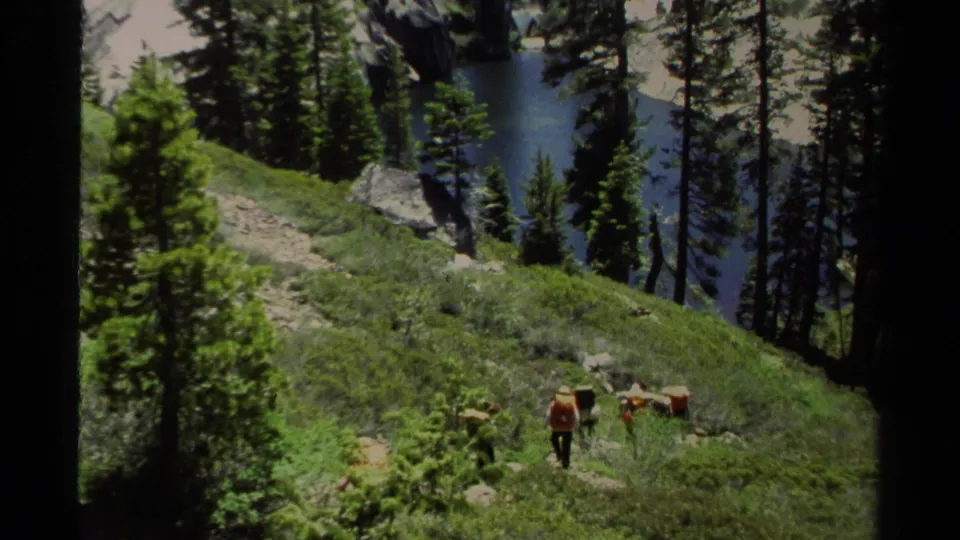 1978 Lake Tahoe home-movie frame showing two backpackers on a hillside trail, with pine trees and blue lake water in the background.