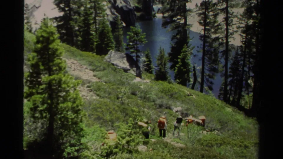1978 Lake Tahoe home-movie frame showing hikers with backpacks on a sunlit pine slope above a blue alpine lake.