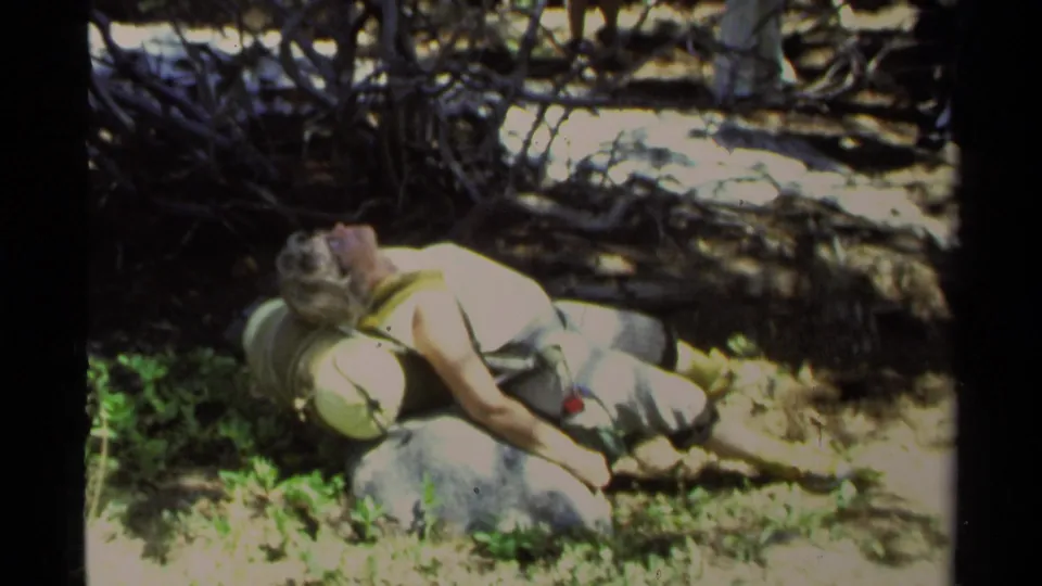 Adult woman in a light-colored dress rests on her back among rocks, plants, and exposed branches in the Lake Tahoe 1978 home-movie clip.