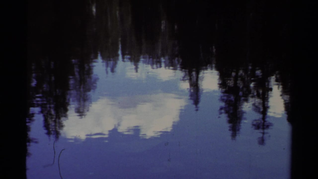 1978 Lake Tahoe 8mm frame showing calm water mirroring dark evergreens, pale clouds, and a snowy shoreline.