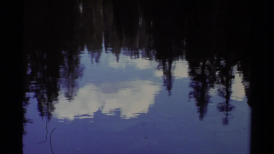 1978 Lake Tahoe 8mm frame showing calm water mirroring dark evergreens, pale clouds, and a snowy shoreline.