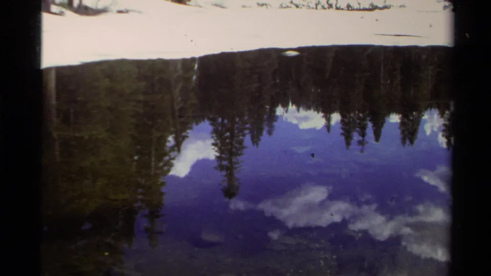 1978 8mm Lake Tahoe view with blue sky, clouds, pines, and snow mirrored on the lake.