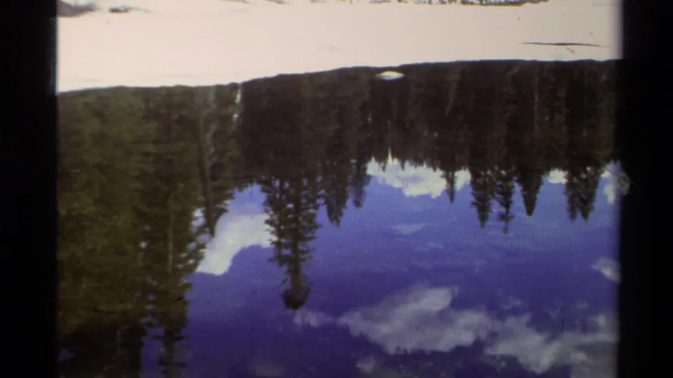 Lake Tahoe winter landscape on 8mm film, with snowy shore, dark pines, and blue cloud reflections in still water.
