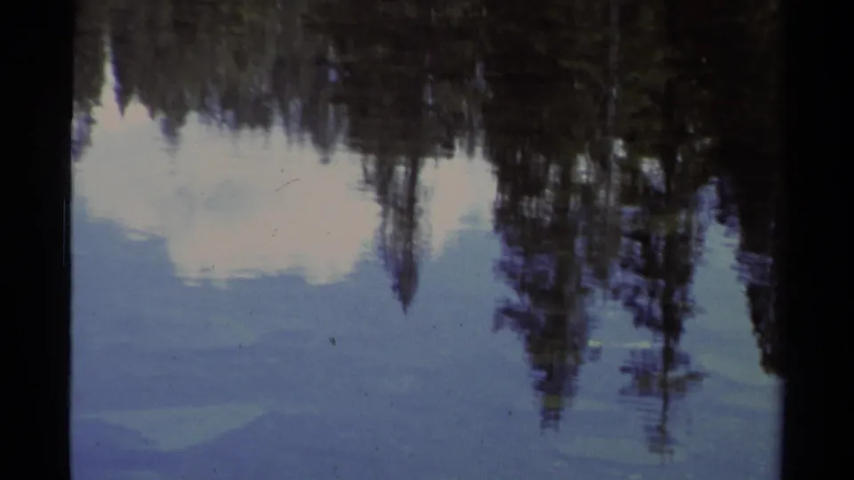 Final Lake Tahoe home-movie frame with the zoom tighter on tree and cloud reflections in calm blue water, with analog film texture visible.