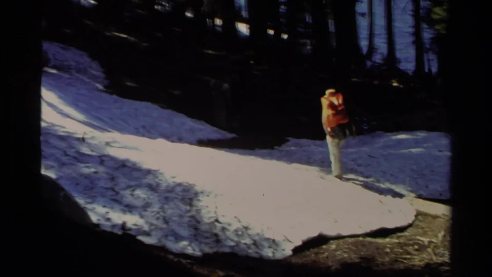 1978 Lake Tahoe frame with a hiker wearing an orange backpack crossing a bright snow patch near shaded pines.