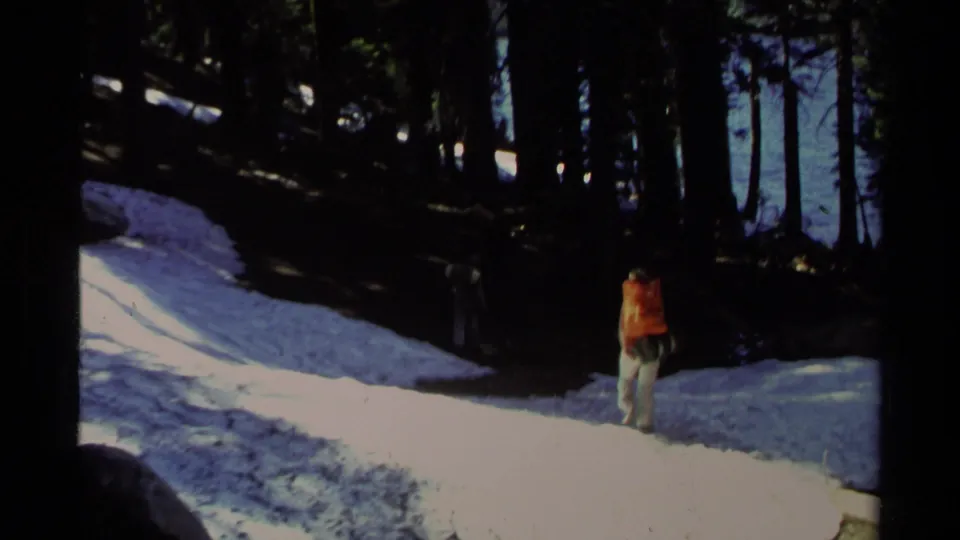 Lake Tahoe 1978 home-movie still with a person in an orange backpack walking away across snow beside shaded forest.