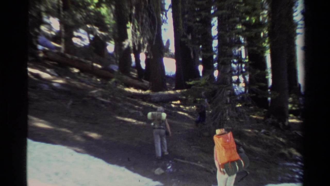 Two hikers with large packs walk along a shaded forest trail with patches of snow nearby.