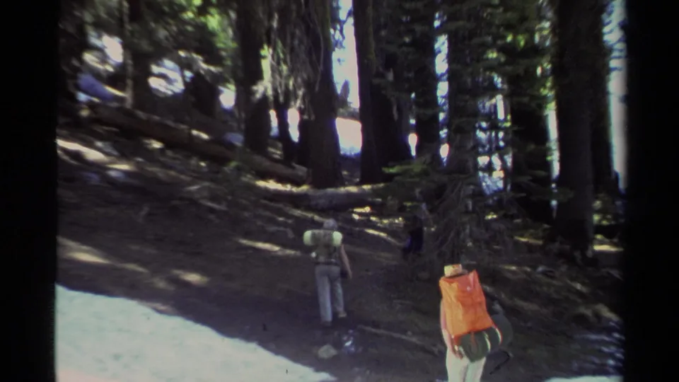 1978 Lake Tahoe home-movie frame showing hikers from behind on a wooded trail with tall conifers, snow patches, and backpacking gear.