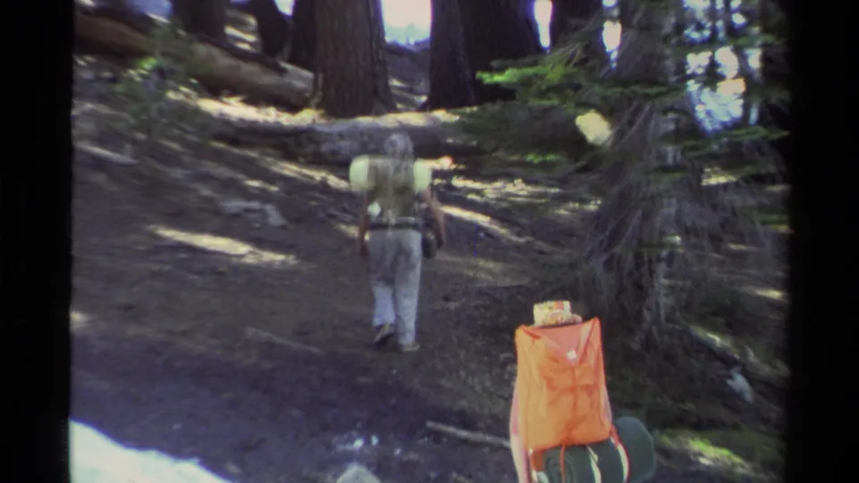 1978 Lake Tahoe hiking footage frame with two backpackers walking away through conifer woods beside lingering snow.