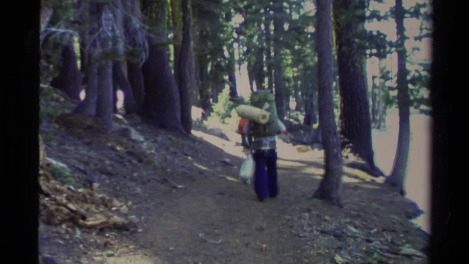 1978 Lake Tahoe home-movie frame showing a backpacker on a conifer-lined trail, with bright light beyond the woods.