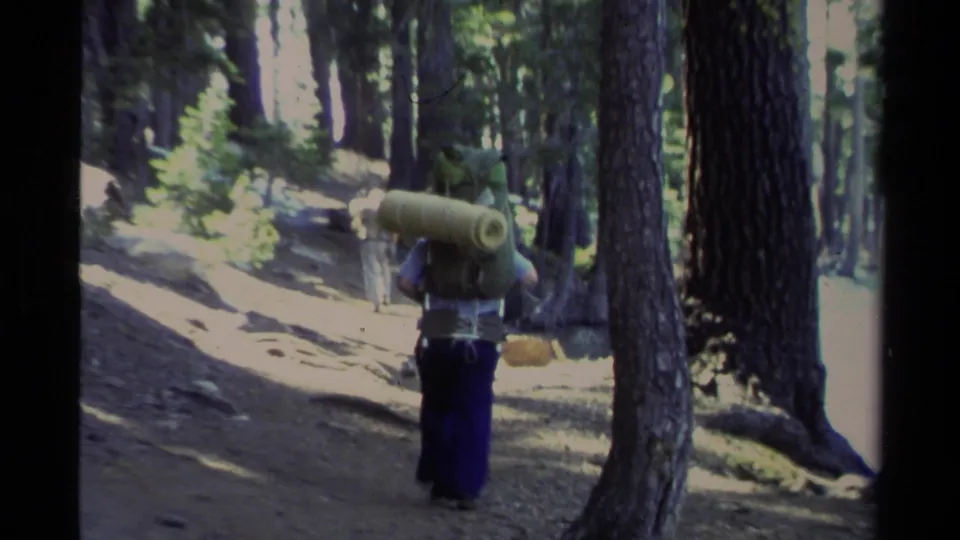 A hiker in blue pants carries a bulky pack and sleeping mat through a shaded Lake Tahoe pine forest in 1978 footage.