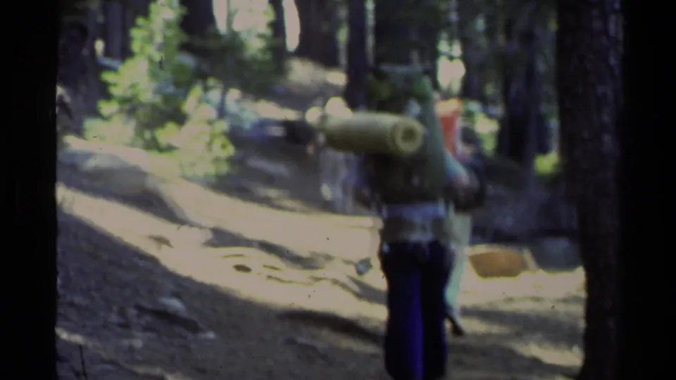 The 8mm frame shows a softly focused backpacker from behind on a Lake Tahoe forest trail, with tall pines and strong sun exposure.