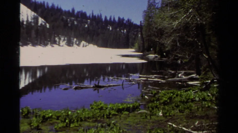 A quiet Lake Tahoe area lake scene shows snow along the far bank, pine-covered slopes and dark water reflections in 1978 home-movie footage.