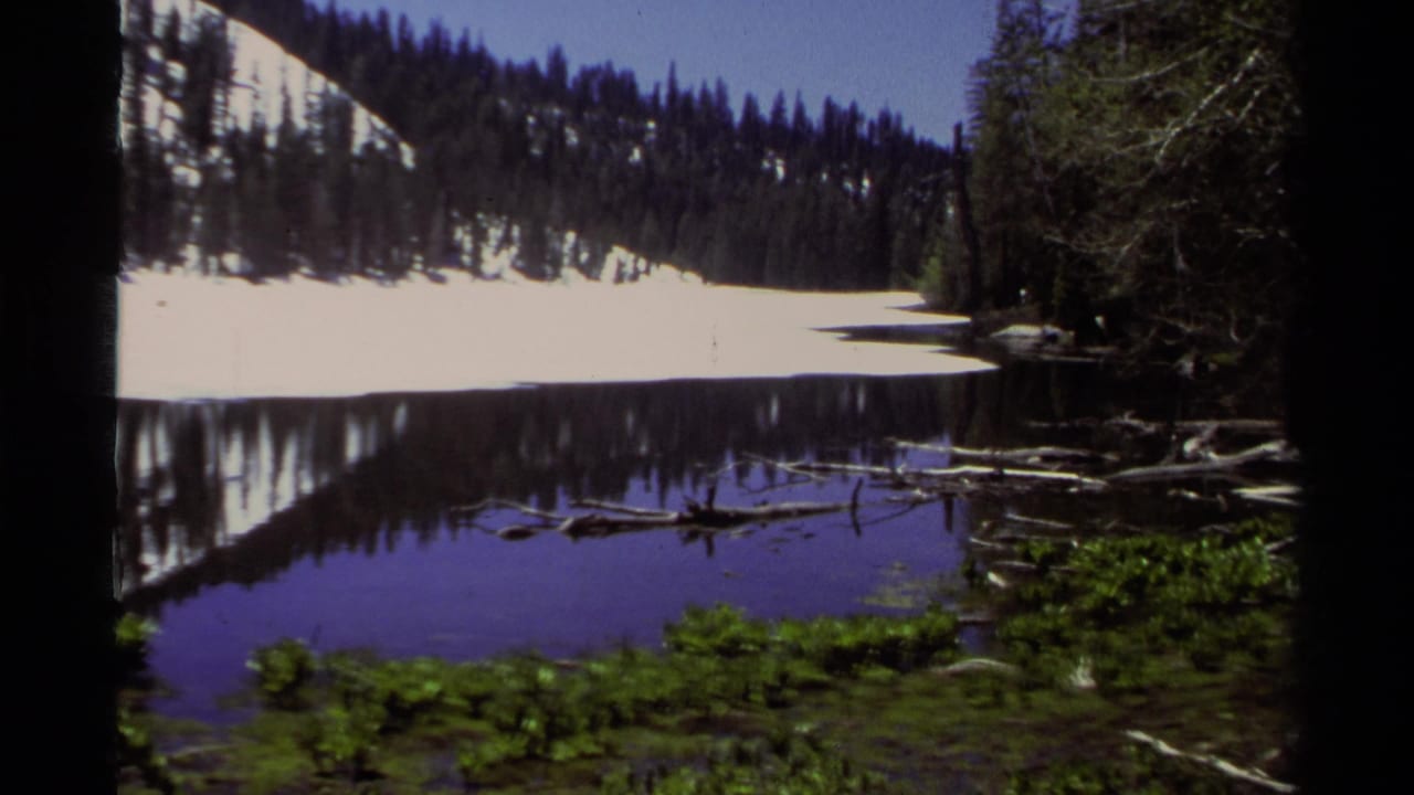 Lake Tahoe home-movie frame from 1978 showing snow on a forested hill reflected in still water, with shoreline vegetation in front.