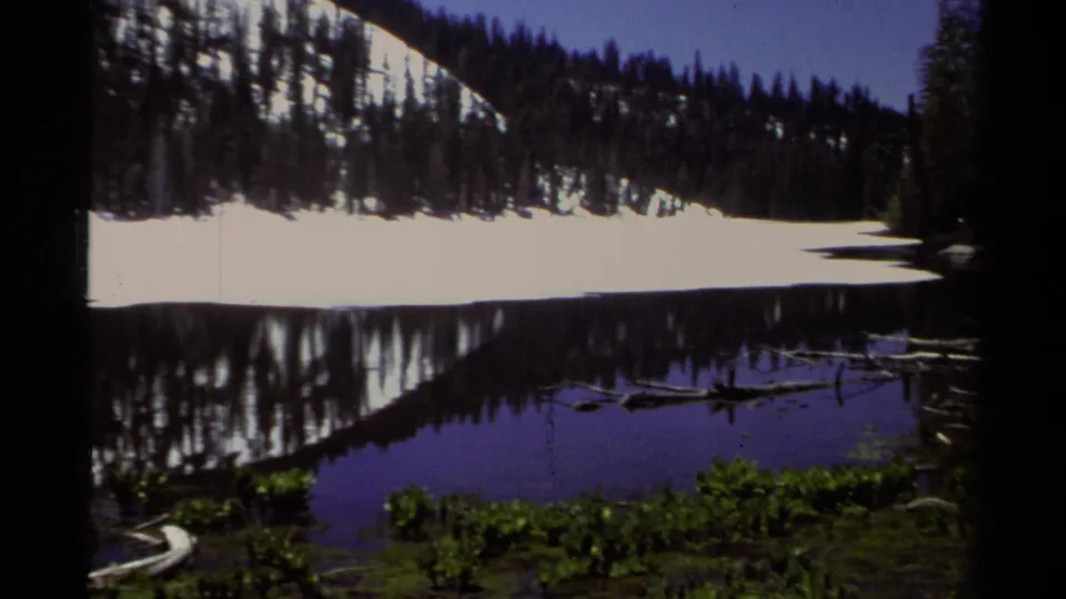 1978 Lake Tahoe 8mm frame with a snowy tree-lined slope reflected in dark blue water and low vegetation near the shore.