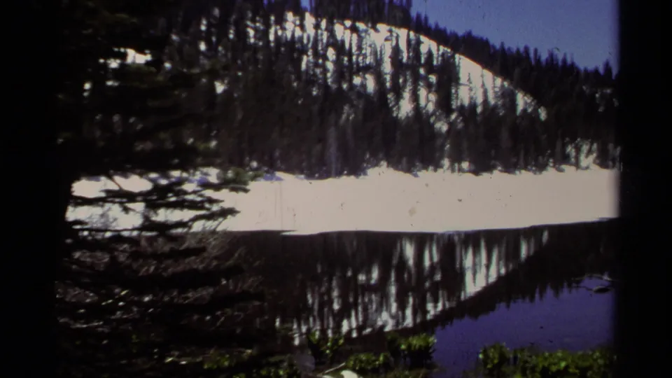 Final 1978 Lake Tahoe frame with shaded evergreen branches at left, a bright snow band, forested slope, and reflected lake water.
