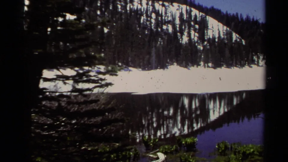 1978 Lake Tahoe home-movie frame showing snow along a wooded slope reflected in calm water beneath dark pine framing.