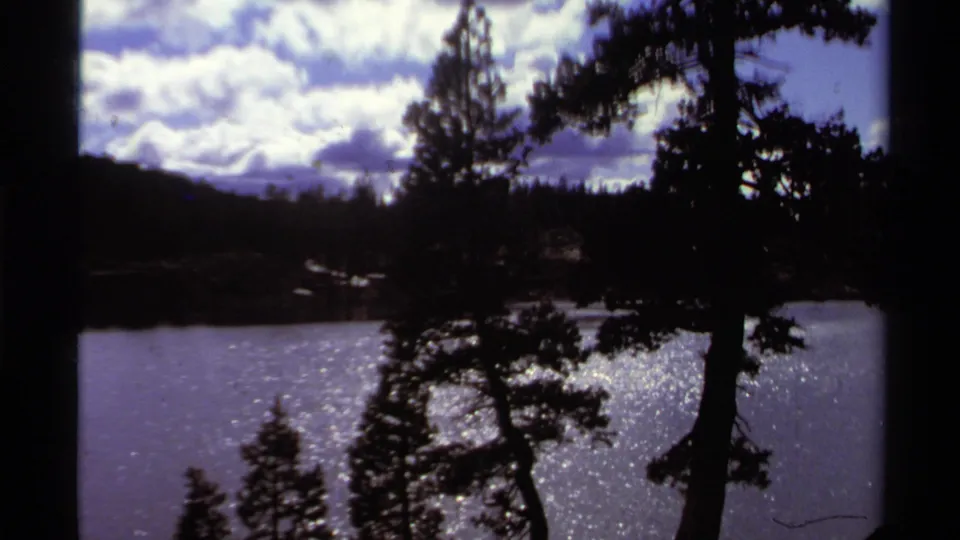 Lake Tahoe home-movie frame from 1978 showing bright cloud cover, a dark treeline, shimmering water, and foreground pines.
