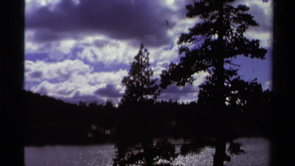 Final 1978 Lake Tahoe frame with large clouds, silhouetted pines, dark wooded ridge, and reflective water below.