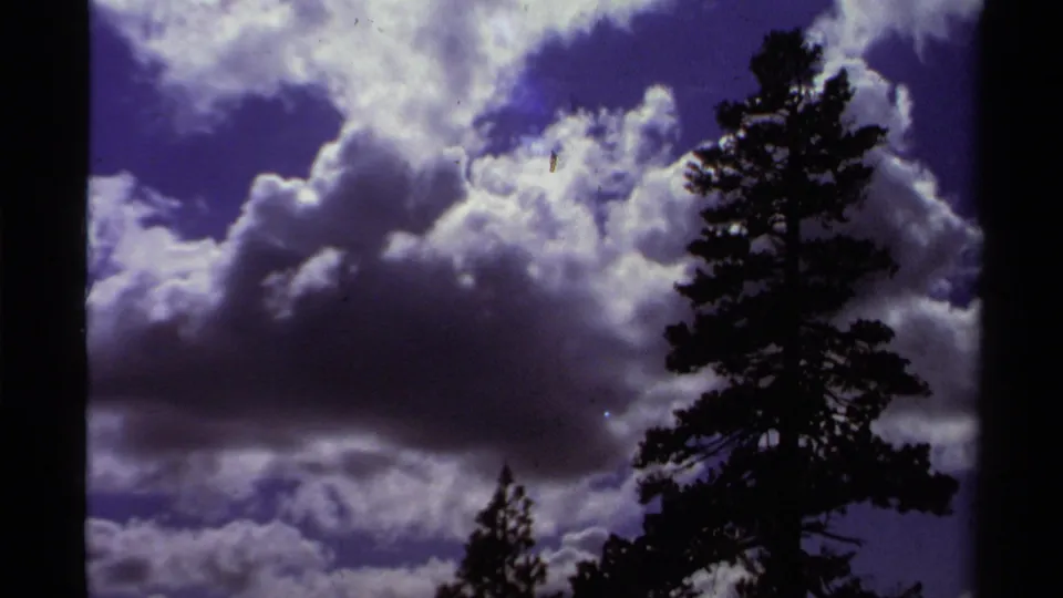 1978 Lake Tahoe 8mm frame showing a tall pine at right against bright layered clouds and saturated blue-purple sky.