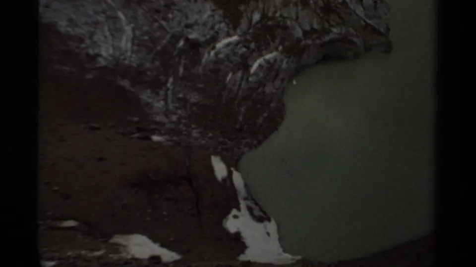 1979 British Columbia 8mm frame showing a rocky lake edge, reddish soil, pale shoreline marks, and green water.