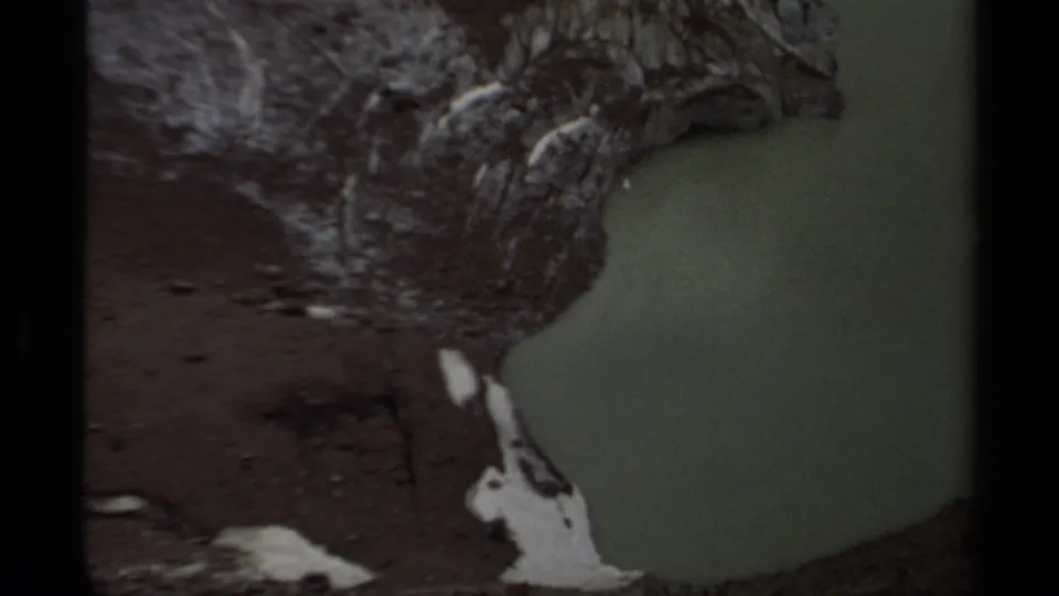 1979 British Columbia 8mm frame of a dim rocky shoreline beside green water, with visible film borders and muted color.