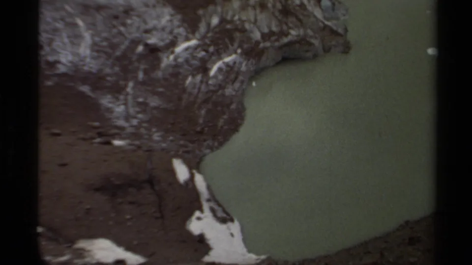 1979 British Columbia 8mm frame showing a murky green pool below dark rocky terrain with patches of snow nearby.