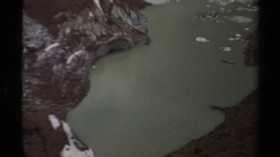 1979 British Columbia 8mm still of opaque green water beside rough volcanic-looking rock and small snow remnants.