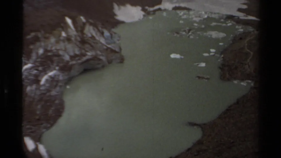 1979 British Columbia 8mm frame showing a broader green pool with rocky banks, snow remnants, and small floating debris or ice.