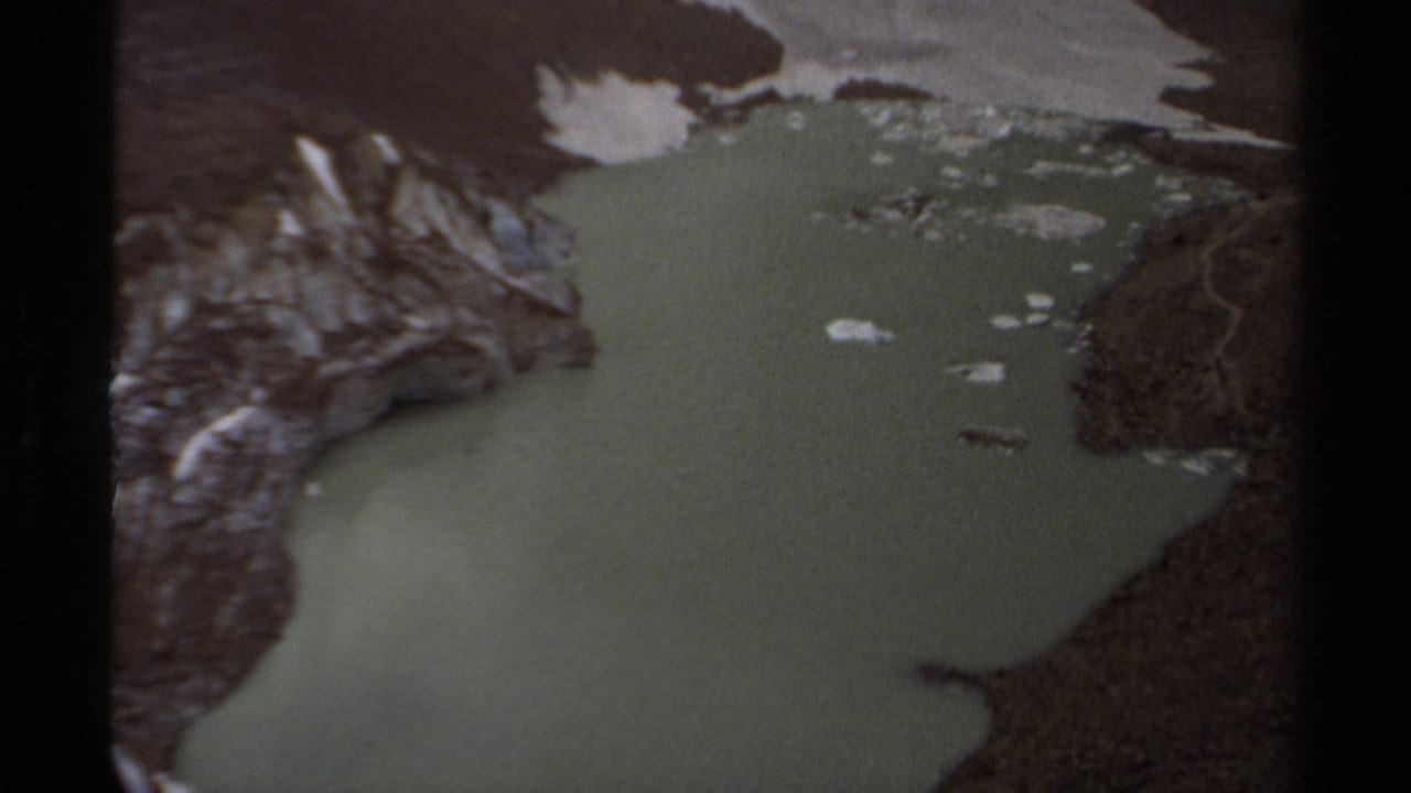 Aerial view of a green alpine lake bordered by rocky slopes and snow patches.