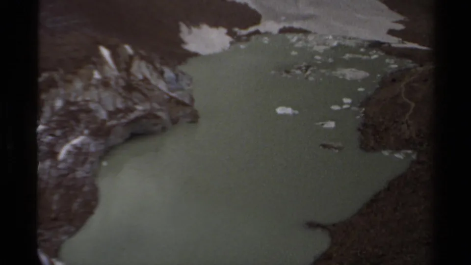 1979 British Columbia 8mm aerial view of a muted green lake below rocky, snow-dusted mountain terrain.