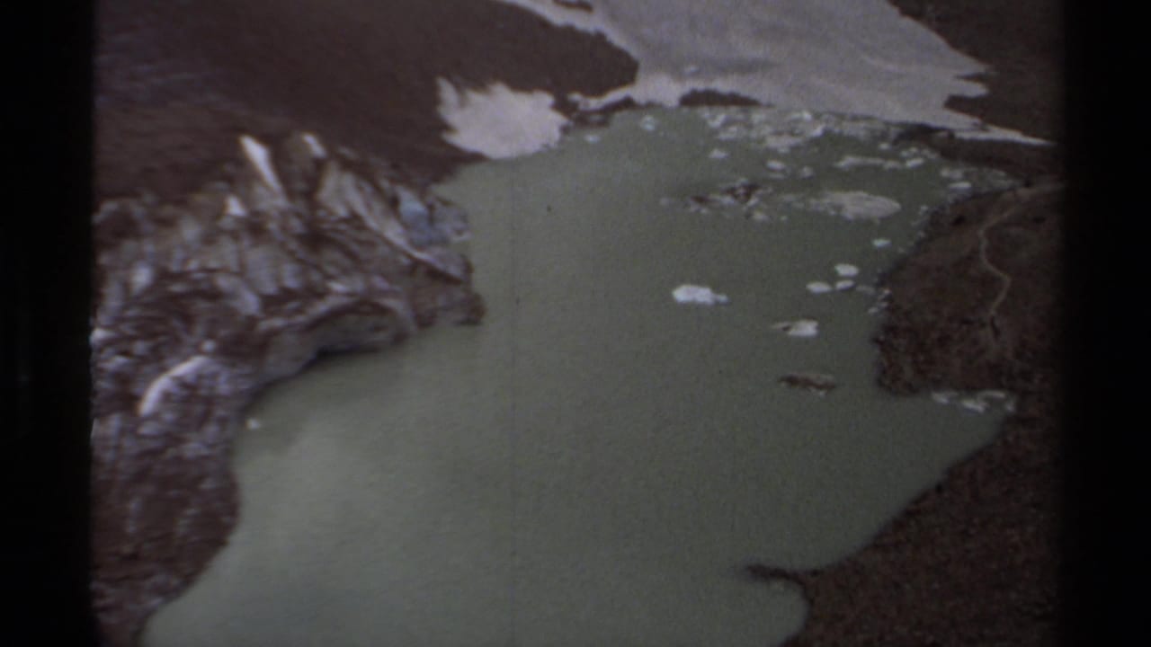 Aerial lake view with snow patches, steep rock faces, and dark shoreline edges.