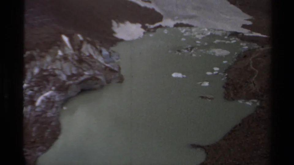 A 1979 8mm aerial frame shows green lake water, snow-streaked rocks, and rugged British Columbia mountain slopes.