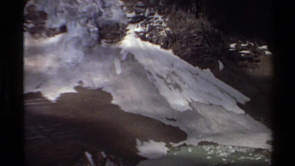 1979 British Columbia aerial frame with a snow-covered slope, exposed rock, and green lake water at the lower edge.