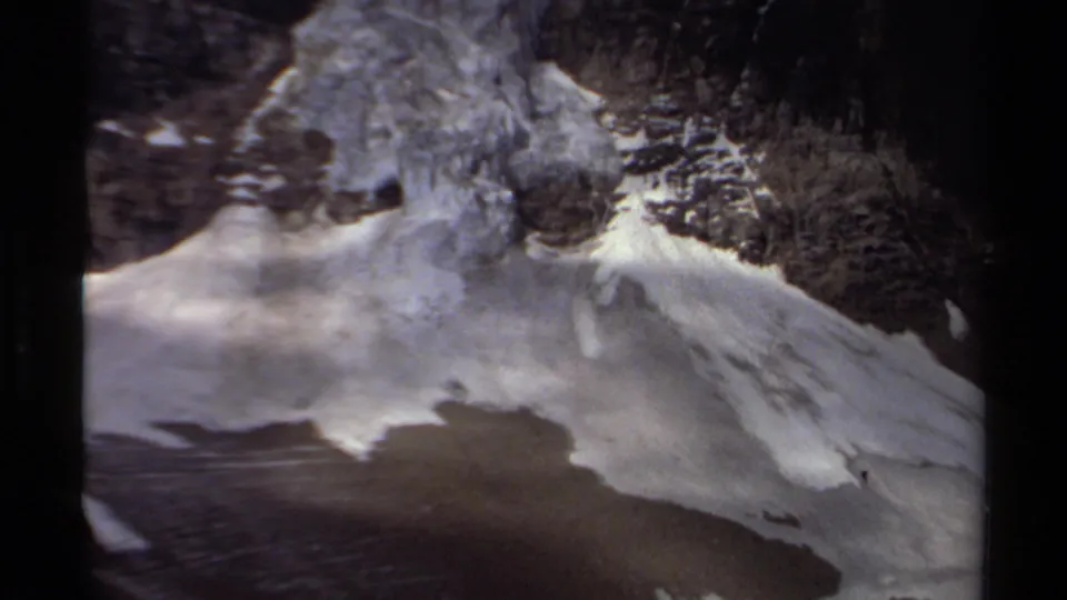 A late-1970s 8mm aerial view in British Columbia shows snow, rock, and a calm alpine lake below.