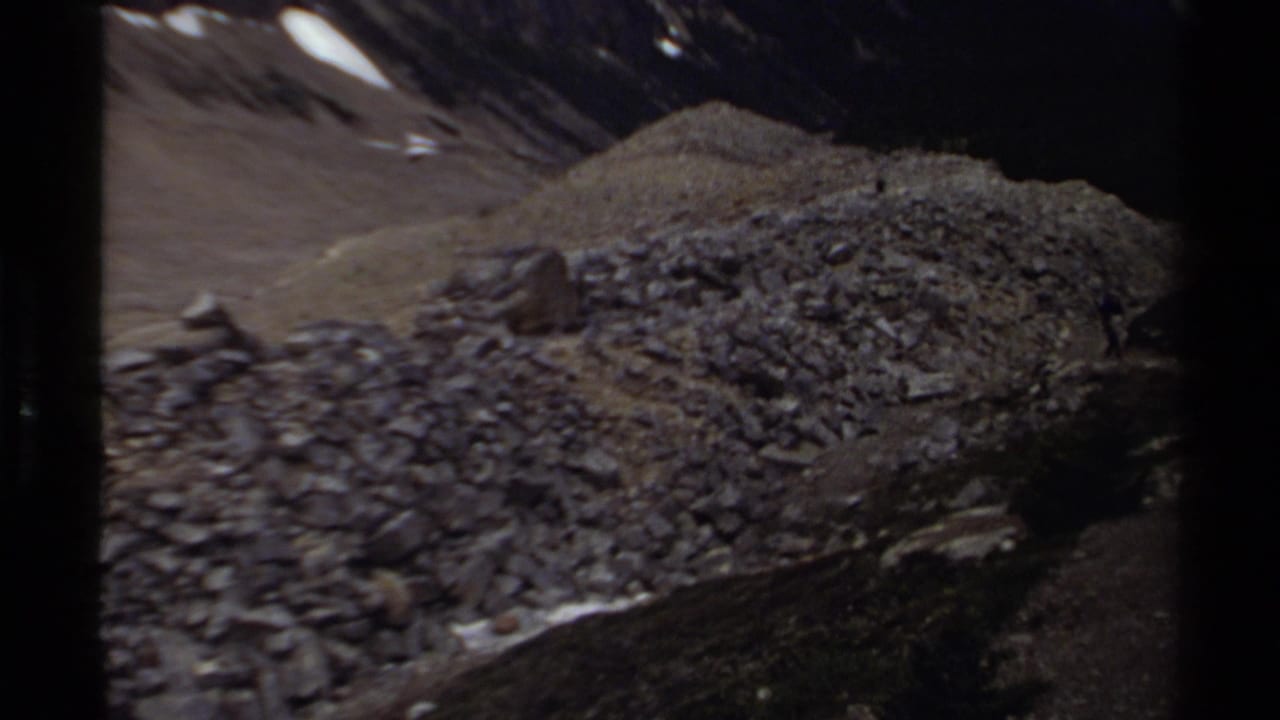 1979 8mm British Columbia mountain view showing a debris-covered slope, small snow patches, and shadowed peaks beyond.