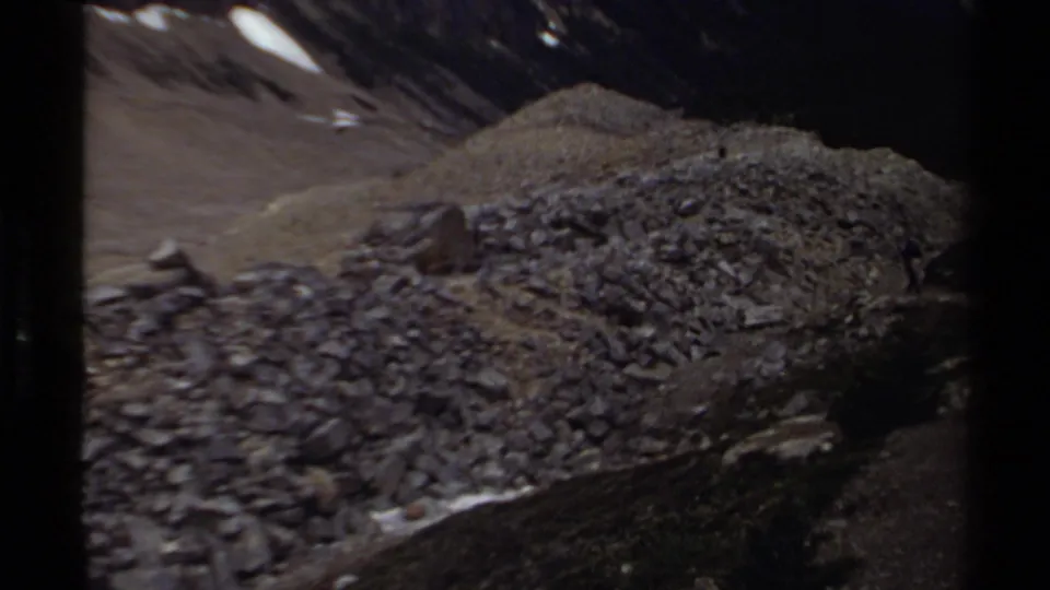 1979 8mm British Columbia mountain view showing a debris-covered slope, small snow patches, and shadowed peaks beyond.