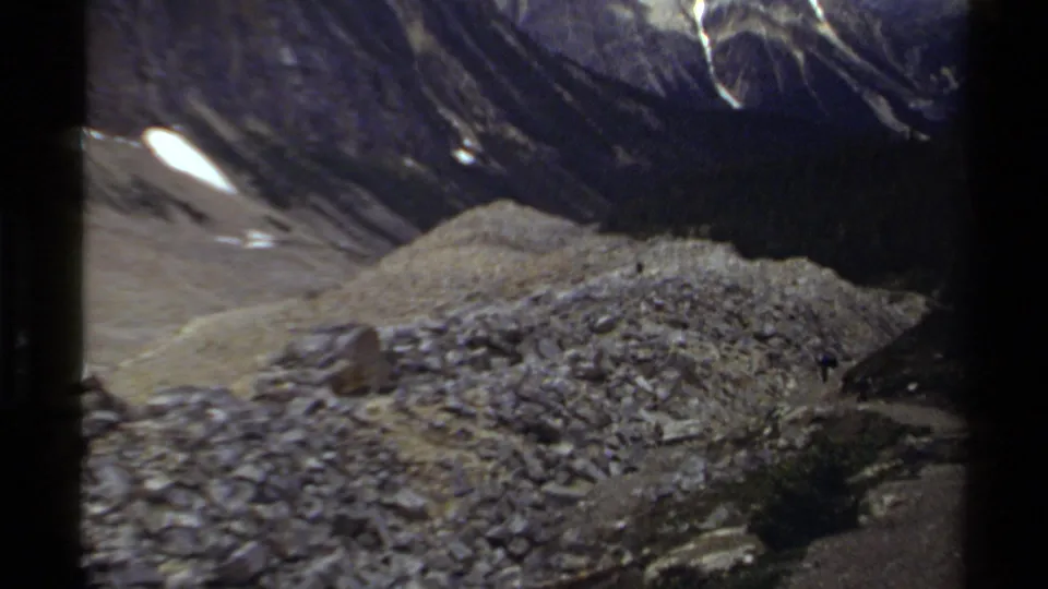 1979 British Columbia home-movie frame of a rocky alpine rise beneath dark mountain slopes and small patches of snow.