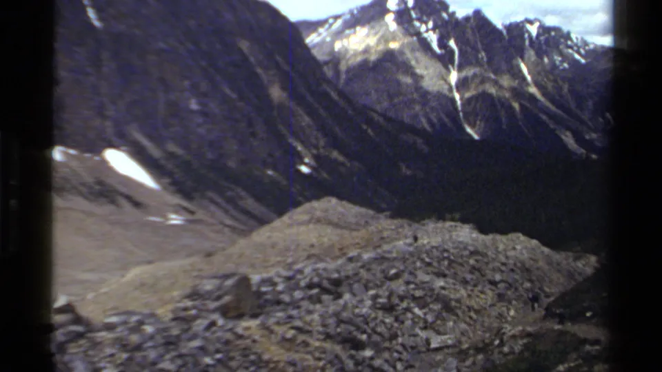 Wider 8mm view of British Columbia alpine terrain, showing a rocky foreground, snow-streaked mountains, and tiny distant people on the slope.